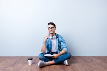 Dreamy student is sitting on the floor at home with crossed legs, thinking, making notes, relaxing with cup of coffee. He is in a casual jeans outfit, glasses