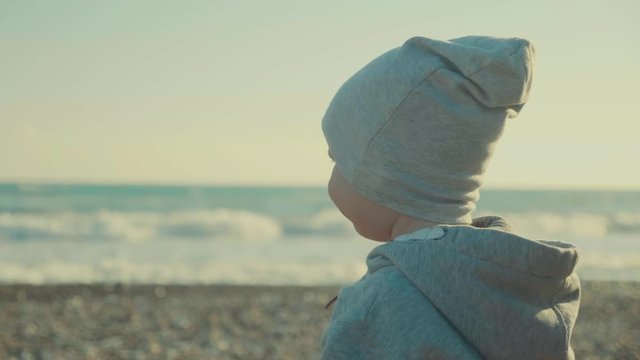 SLOW MO little cute boy on an empty beach looking at the amazing sea with foam waves, in the background a horizon line. Child walks on the seashore