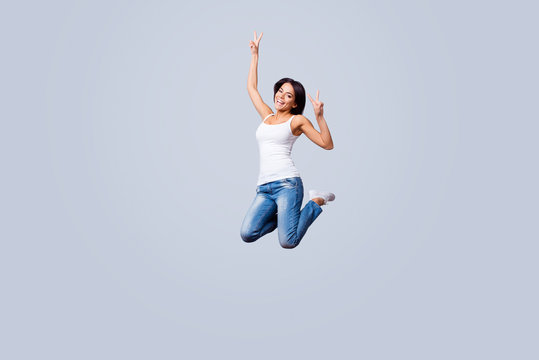 Go crazy! Happy young brunette latin girl is jumping and fooling around. She is in casual outfit, showing v sign, isolated on a pure light background