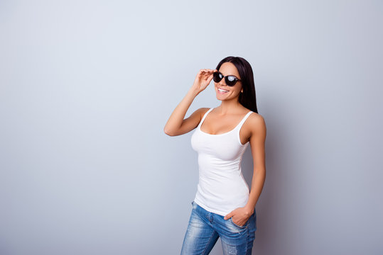 Young cute latin american lady with beaming smile in stylish spectacles is standing on the light blue background. She is full of dreams and fantasies, wearing jeans and white singlet