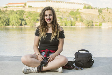 Portrait of a beautiful female photographer with a professional camera. Woman photographer with a professional camera and bag. Beautiful day by the river. Lens flare in the background.