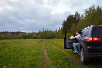 Fototapeta premium Young man sitting in his car with the door open in a beautiful landscape. Off-road travel on vacation, enjoy the freedom