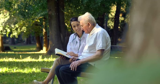 A nanny, a nurse, caring for the elderly a girl (woman) and grandfather sit on a paperback and read a book, in the park. Concept boarding house, sanatorium, house for the elderly, help for the elderly