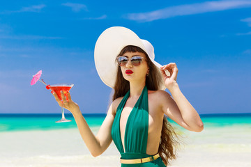 Beautiful brunette in a straw hat with a martini glass on the shore of a tropical beach. Summer vacation concept.