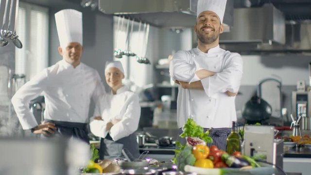 Famous Chef of a Big Restaurant Crosses Arms and Smiles in a Modern Kitchen. His Staff in Smiling in the Background. Shot on RED EPIC-W 8K Helium Cinema Camera.