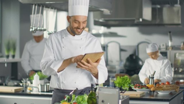  Famous Chef Uses Tablet Computer for Recipes While Working in a Modern Kitchen. His Help Work in the Background.Shot on RED EPIC-W 8K Helium Cinema Camera.