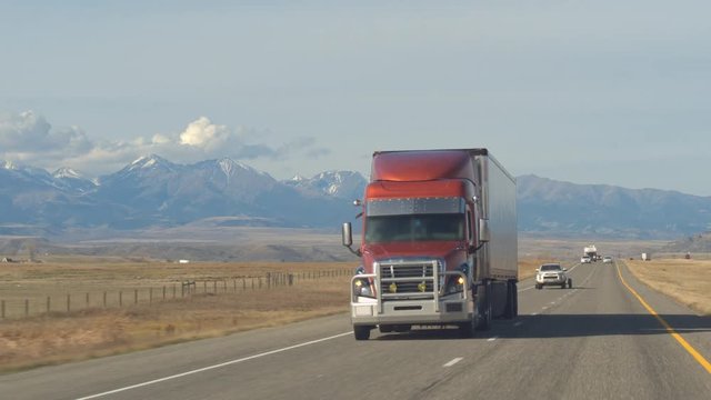 CLOSE UP: Freight semi truck transporting goods driving along the multiple lane highway past pasture fields and farmlands. Lorry on sunny day with snow hooded Rocky Mountain Range in the backgrounds