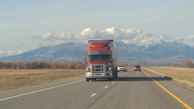 CLOSE UP: Freight semi truck transporting goods driving along the multiple lane highway past pasture fields and farmlands. Lorry trucking load on sunny day with Rocky Mountain Range in the backgrounds