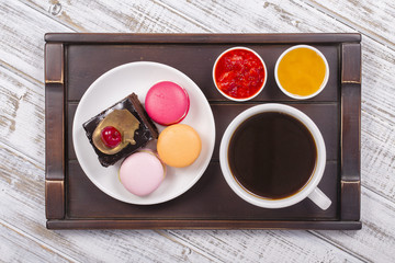 Cup of coffee, honey, strawberry jam, piece chocolate cake and macaron cakes on tray on white wooden table. Lifestyle concept.