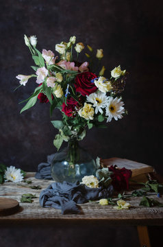vase with a bouquet of assorted flowers on a wooden table with old books, grey cloth, petals on a brown background
