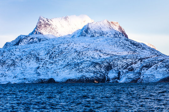 Huge Sermitsiaq mountain covered in snow with blue sea and small fidhing boat in the foreground, nearby Nuuk city, Greenland