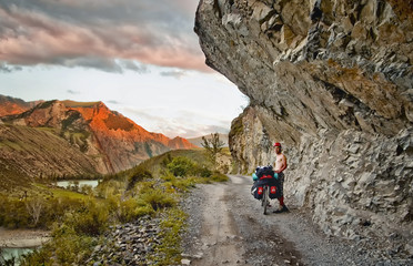 Velopohd in Altai, Russia, summer. Male cyclist standing on a mountain road in the cliff. In the background, mountains and sky. The road is carved into the rock on a steep cliff above the river.