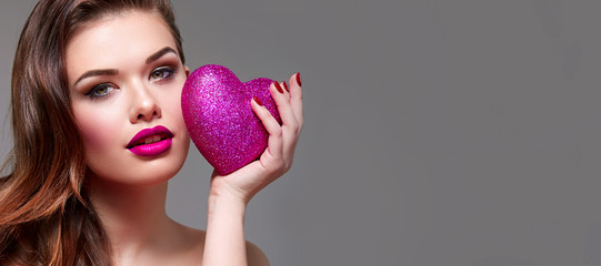 Obraz premium Beautiful young attractive girl in studio on a gray background holds in hands a large shiny pink heart. Romance, love. Makeup - pink lips, golden shadows. Hairstyle is a Hollywood wave.