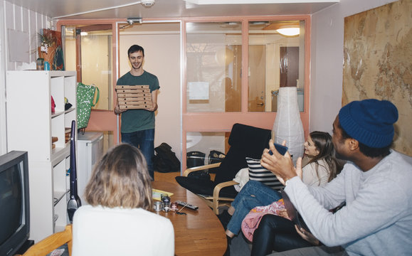 Happy young roommates sitting with man carrying stack of pizza boxes at college dorm