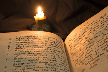 Open book with candle soft light pouring on text. Reading of opened book educate reader. Ancient Bible text reading on black background. Russian bible. Old slavonic text and letters