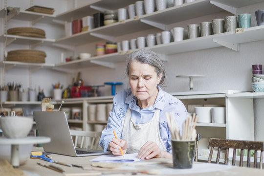 Senior female potter writing on paper while sitting with laptop at table in workshop - Powered by Adobe