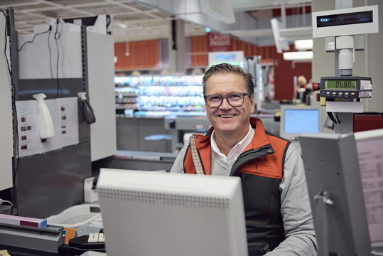 Portrait of smiling mature cashier sitting at checkout counter in supermarket