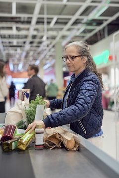 Mature woman standing with groceries and bag at checkout counter in supermarket