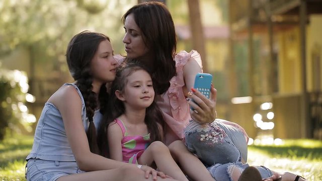 Mom with two daughters in the nature sitting on the grass and making a selfie with a phone