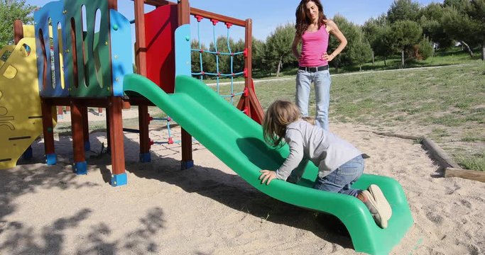 Three years old blonde child playing trying to climb on green plastic small slide, next to woman mother. Outdoor playground, in public Park Valdebebas, Madrid Spain

