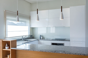 Modern kitchen furniture design in light interior with wood accents. Kitchen peninsula in the room. Stone surface. Gloss facades. Finished project. White pendant light. Sink, window, blinds. Glass.