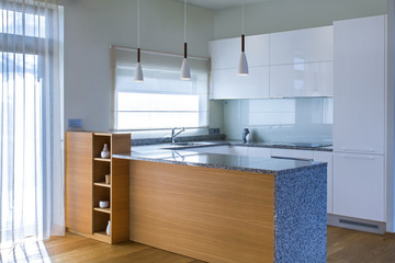 Modern kitchen furniture design in light interior. Walnut veneer. Kitchen peninsula in the room. Stone surface. Gloss facades. Finished project. White pendant light. Sink, window, blinds. Glass.