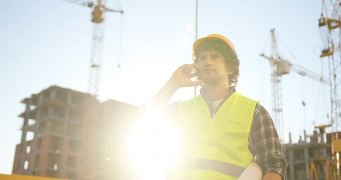 Beautiful builder with black curly hair in yellow helmet and green vest talking on cell phone with friend, smiling on unfinished construction background.