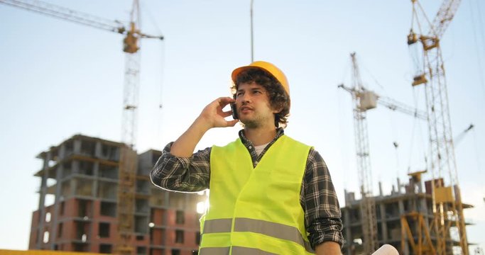 Young builder with black curly hair in yellow helmet talking on mobile phone with friend, smiling on unfinished construction background.