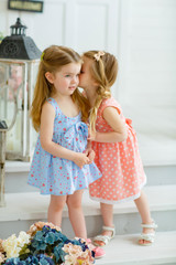 Two little girl friends are whispering, telling secrets. They stand on the white steps at the entrance of the house.