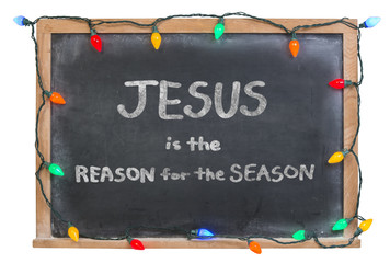 Fototapeta na wymiar Jesus is the reason for the season written in white chalk on a black chalkboard surrounded with festive colorful lights isolated on white