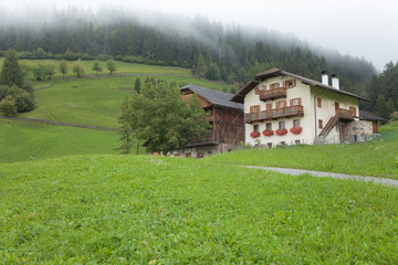 Traditional houses in a cloudy mornig surrounded by green pasture next to the little town of St. Magdalena in Val di Funes, no people around