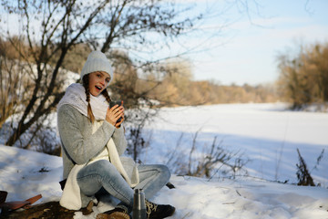 Cheerful young woman resting on nature, in a snowy forest near a river, holds a thermos mug with happiness, a healthy lifestyle