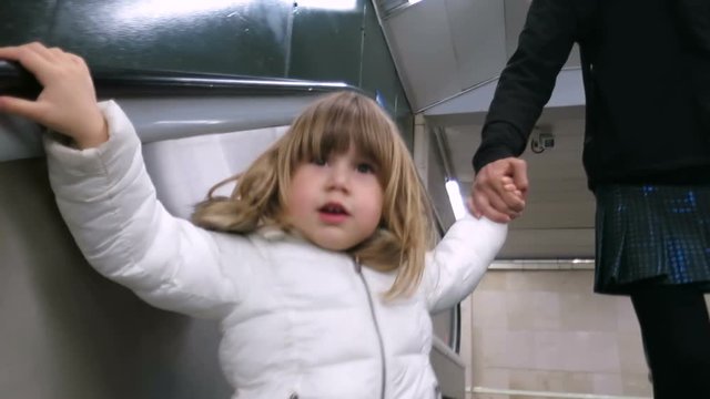 three years old blonde child with white coat holding mother’s hand walking in mechanic stairs of subway or underground station of Madrid city
