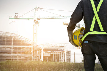 Rear view Asian  male site contractor engineer with hard hat holding blue print paper inspecting at construction site, crane with golden sunlight at the background.