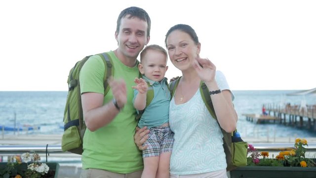 A young family with a toddler looking at the camera and smiling. Everyone has backpacks on their backs. The sea and the beach behind.