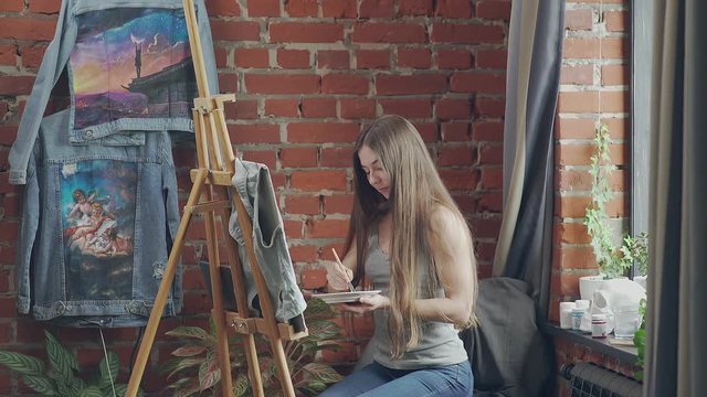 Beautiful long-haired woman painter paints on canvas of jeans. Young stylish woman with long hair draws at the easel in the loft. Loft space: a stylish interior in an art studio.