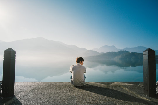 Lonely Asian man sitting alone on a public pier on a clear lake in an early morning. The lake is very clear and can see the reflection of all mountains. The sun shine on to the lonely man / guy. 