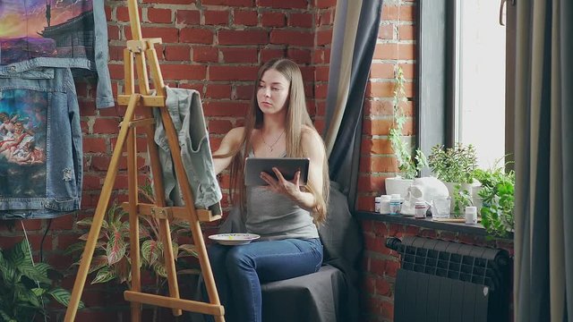 Long-haired girl paints with oil colors on easel in workshop interior. Young stylish woman with long hair draws at the easel in the loft. Loft space: a stylish interior in an art studio.
