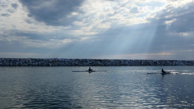 Young single scull rowing competitors paddle on the tranquil lake. Beautiful beams of the sunshine filter through clouds