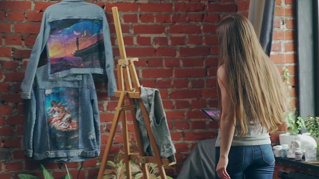 Long-haired girl paints with oil colors on easel in workshop interior. Beautiful long-haired woman painter paints on canvas of jeans. Artist go to easel for drawing, holding a sketch on the tablet.