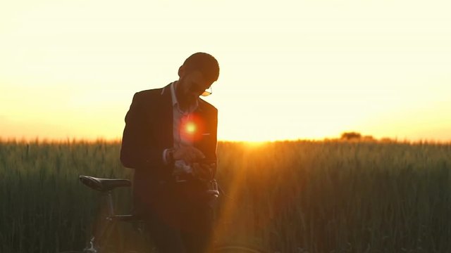 Man in suite and glasses work by selphone in field with bicycle dawn slow motion