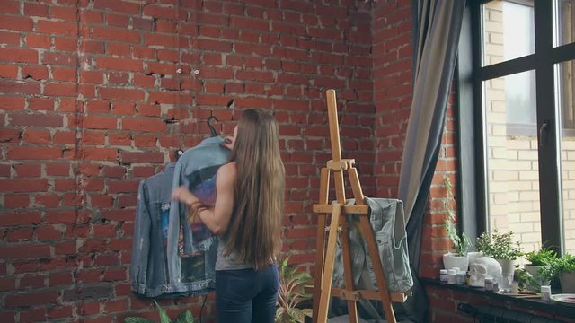 Long-haired and slender woman artist finished work - drawing on a denim jacket. The artist finished drawing on a denim fabric and hangs a thing on the wall. Creative studio in loft style