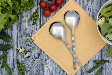Fresh salad with leaves, tomatoes, cucumber and quail eggs, salad spoon and fork, napkin on grey wooden table