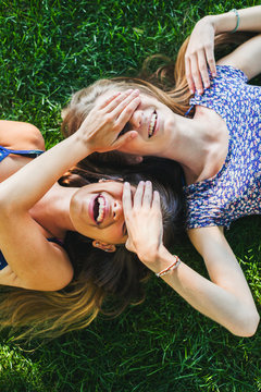 Young female friends covering the eyes of each other whilst lying on the grass