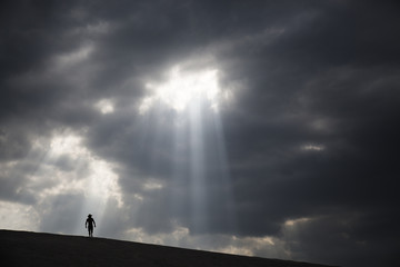 A man walking across sand dunes as a ray of light shines through the clouds