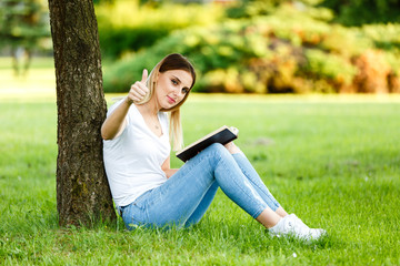 Positive young girl under the three showing thumbs up and reading the book