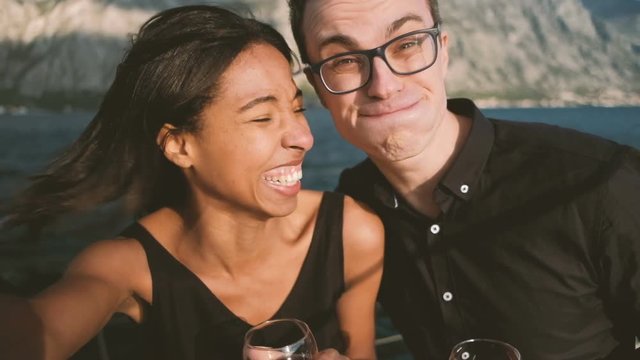 Young man and woman cheerfully doing selfie on ship in open air. Couple in love with glasses of red wine in their hands laughs cheerily, fools around and make faces to camera taking photo with help of
