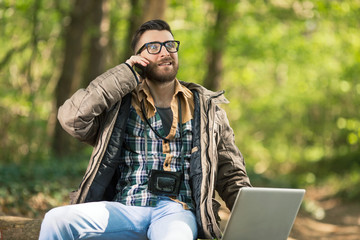 Portrait of busy young man using laptop, mobile phone and camera in the woods