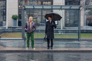 Two men stand at the bus stop, one of them is sad, another is cheerful