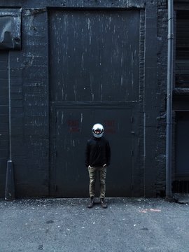 young male standing along in front of grey wall in alley way wearing motorcycle helmet